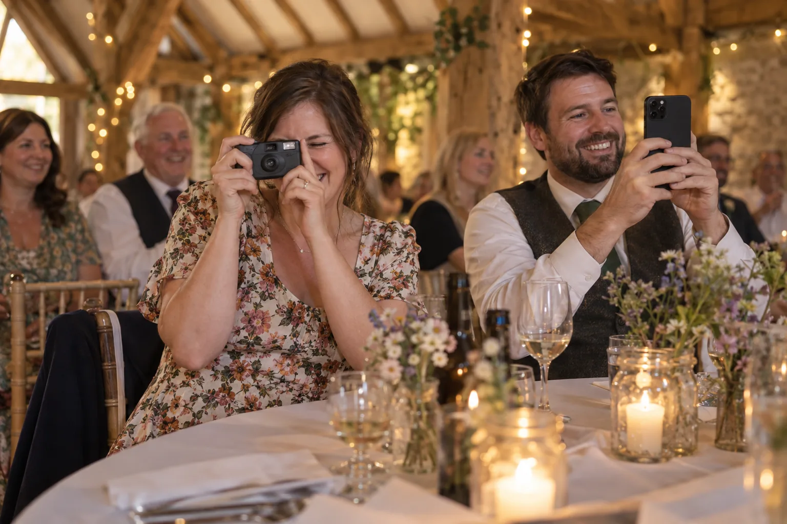 A wedding guest in a barn holds a disposable camera to her eye while the man beside her takes the same photo on his phone