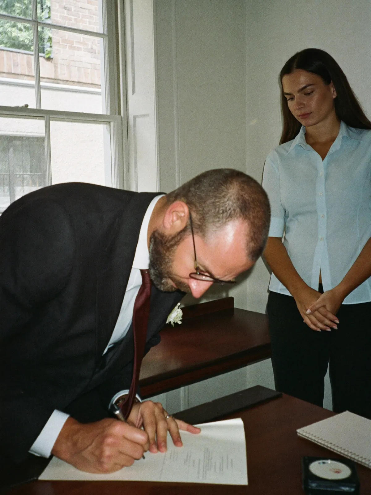 Two witnesses signing the register, heads bent together over the desk